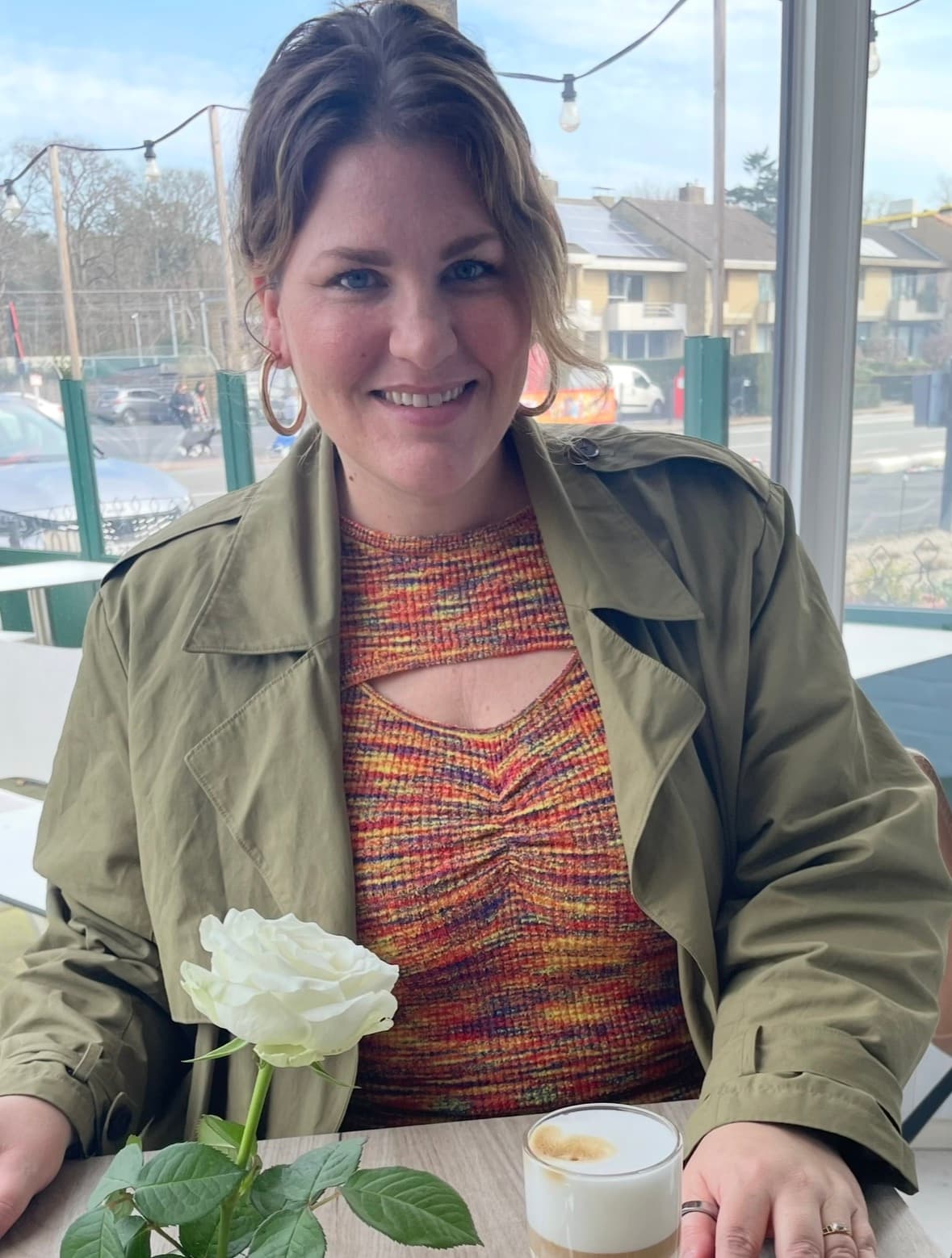 Kathleen Dengerink smiling at a café table, wearing an olive jacket over a multicolored knit top, with a white rose in front of her.